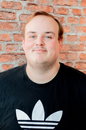 Tim-Lucas Bay Foto zeigt einen Mitarbeiter mit schwarzen T-Shirt und weißem Adidas Aufdruck, vor roter Backsteinwand