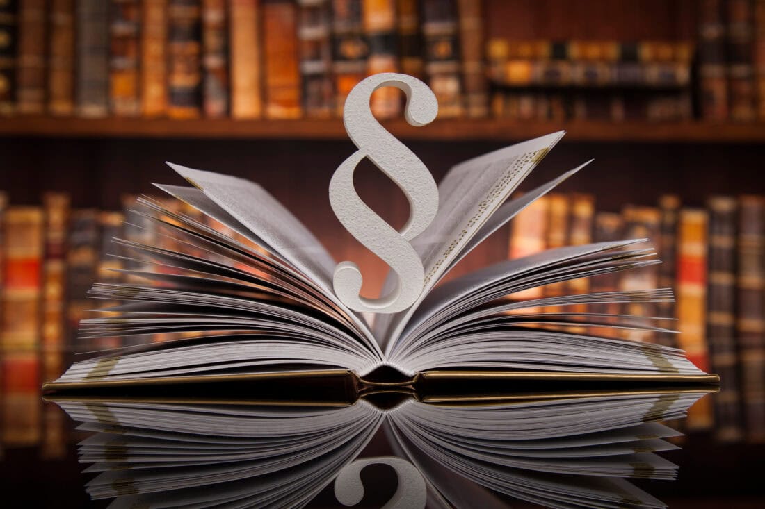 Offenes Buch mit großem Paragraphenzeichen vor einer Bibliothek.